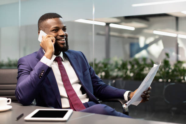 Happy african american businessman sitting at workdesk with modern digital tablet with blank screen on at office, having phone call with business partner, holding papers, panorama with copy space